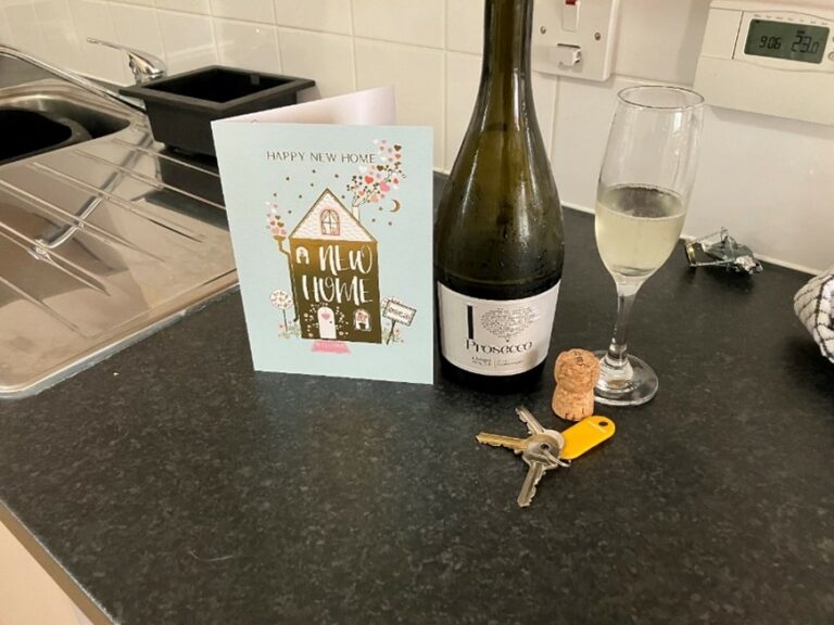 There are items on a table including a bottle of Prosecco, keys, and a card that reads 'happy new home'.