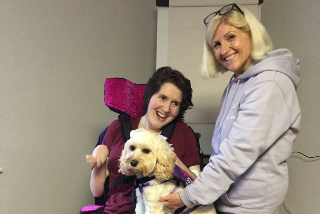 Patient in a wheelchair with Lily, a fluffy white dog on her lap. Jane who is Lily's owner is stood next to the patient.